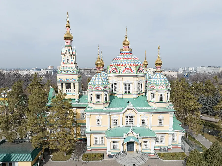 A pastel-coloured wooden cathedral with a tall bell tower on the left and several domes with patterned roofs, seen from above among trees