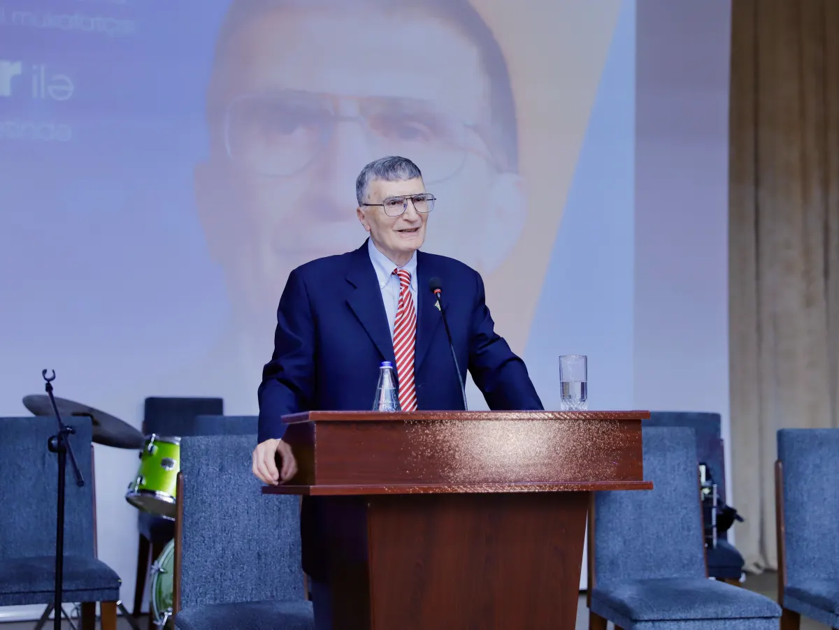عزيز سانجار، رجل متقدم في السن يرتدي بدلة كحلية وربطة عنق حمراء، يتحدث من خلف منبر خشبي على مسرح فيه كراسٍ زرقاء، وخلفه صورة شخصية كبيرة له معروضة على الشاشة