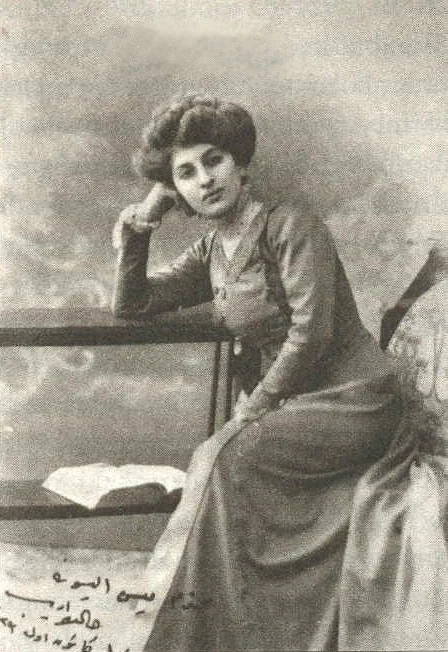 Eine junge Frau mit hochgestecktem Haar, taillierter Jacke und langem Rock sitzt mit einem Arm auf einem Geländer; neben ihr liegt ein aufgeschlagenes Buch. Links unten stehen einige handschriftliche Zeilen in osmanischer Schrift