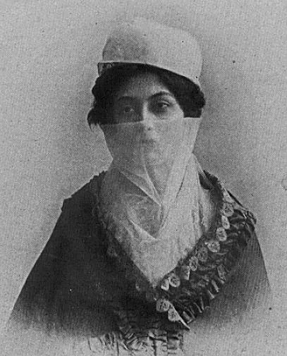 Eine Frau mit einem kleinen runden Hut, einem hellen Schleier über der unteren Gesichtshälfte und einem dunklen Umhang mit verzierter Kante auf einem alten Abzug