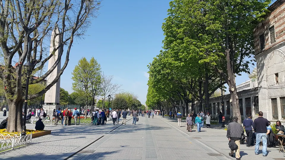 Ein weiter gepflasterter Platz im Sonnenschein mit einem Obelisken links, kahlen und belaubten Bäumen sowie vielen Menschen, die zu Fuß unterwegs sind