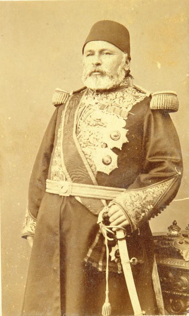 Sepia photograph of H&uuml;seyin Avni Paşa, a stout man with a white beard, in a fez and an embroidered uniform with fringed epaulettes, a sash, a belt and two star-shaped decorations, his hand on a sabre