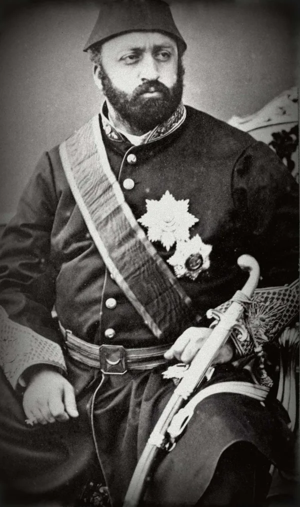 Black-and-white photograph of Abd&uuml;laziz, a heavily built man with a dark beard, in a fez and a dark buttoned coat with a broad sash, two star-shaped decorations and a sabre across his knees