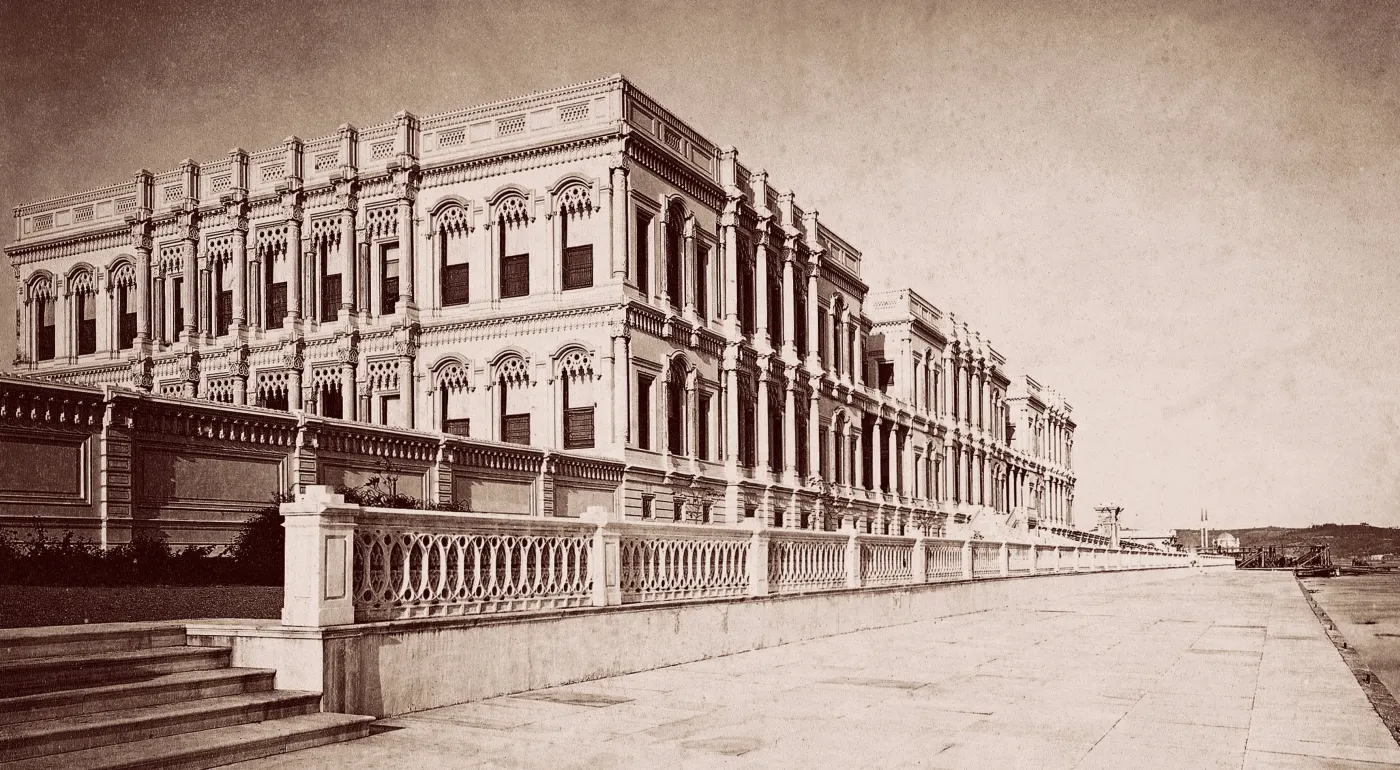 Sepia photograph of a long, three-storey palace of pale stone with rows of arched windows, seen from a stone quay with a balustrade, and a hill in the distance