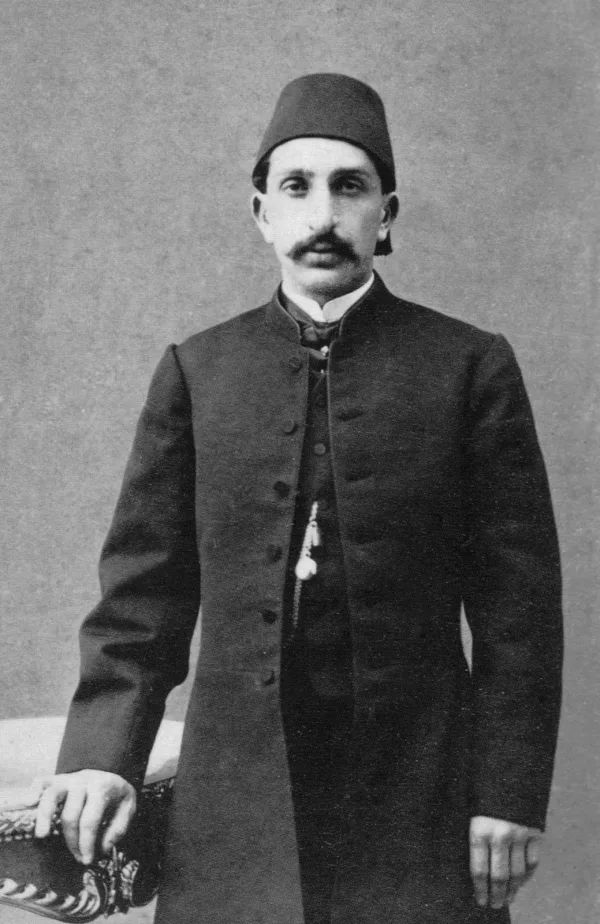 Photograph of the young Abd&uuml;lhamid II, standing with a moustache, in a fez and a long dark coat, one hand resting on a carved chair