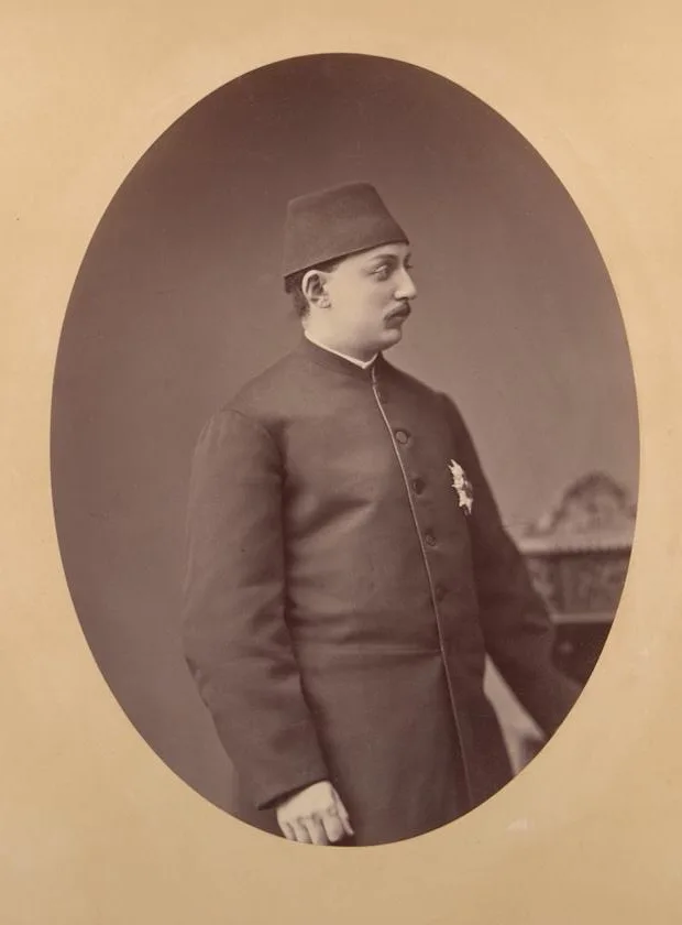 Photograph of a young man with a moustache in a fez and a long dark buttoned coat with a star-shaped decoration, seen in profile and facing right, inside an oval frame