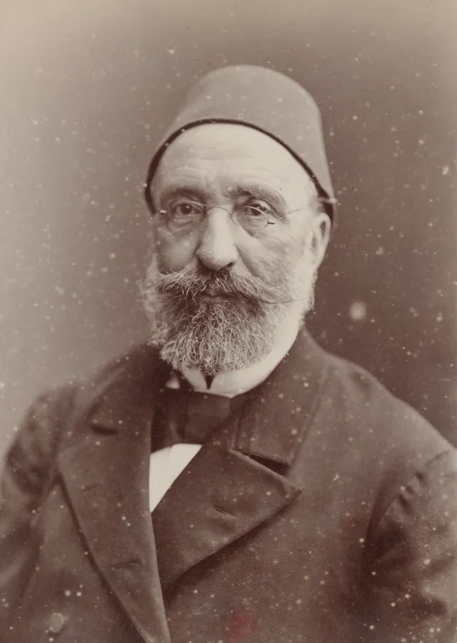 Studio portrait photograph of Midhat Paşa: an elderly man with round glasses and a full grey beard, in a fez and a dark coat with a bow tie