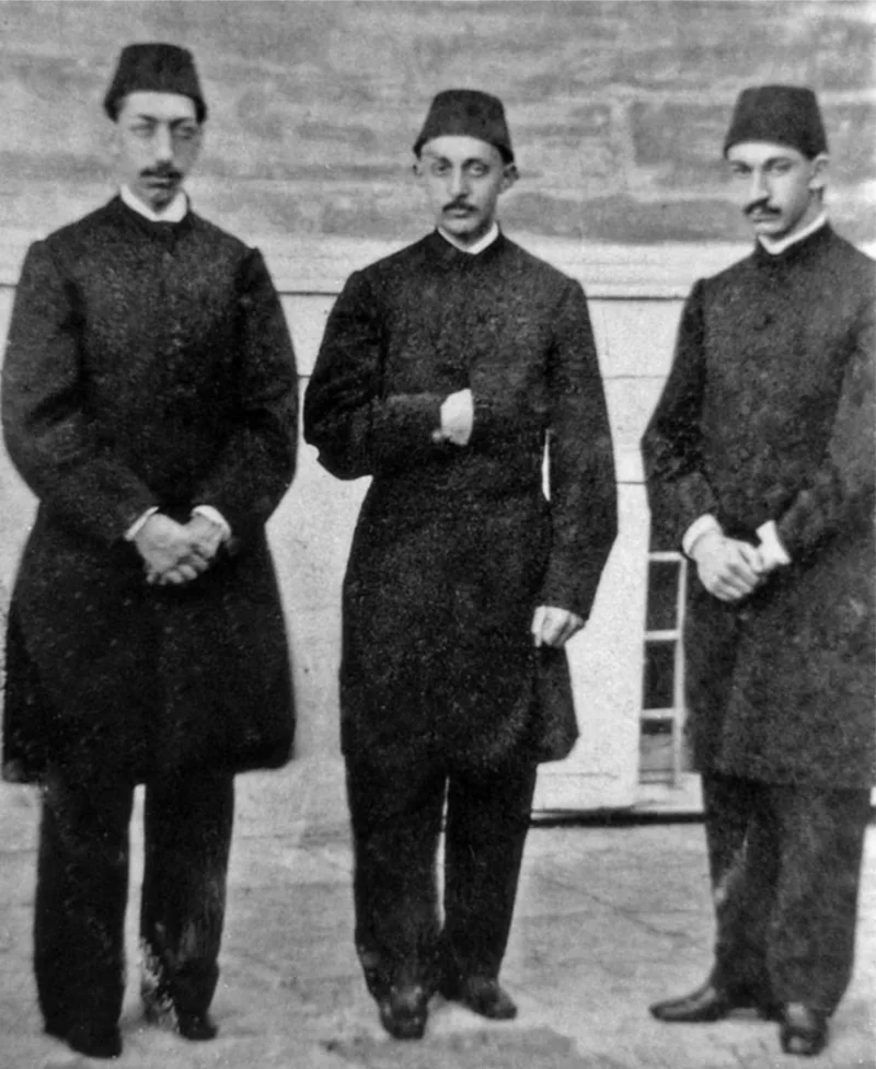 Black-and-white photograph of three young men with moustaches in fezzes and long dark coats, standing side by side in front of a wall; the one on the left wears round glasses