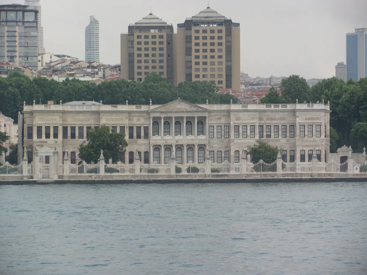 A long pale stone palace building with a pediment in the middle and tall arched windows, behind a low white fence on the Bosphorus shore, seen from the water in daylight; tall modern buildings rise behind the trees