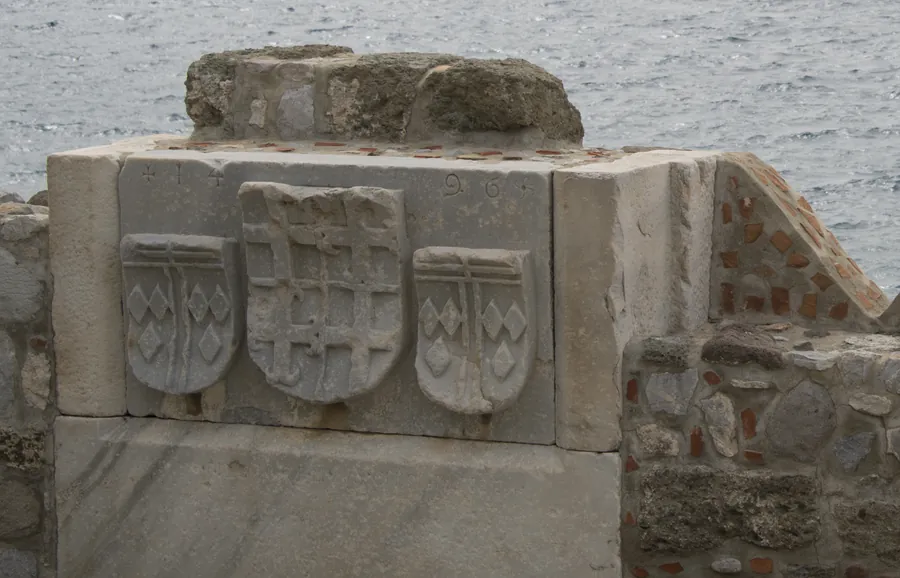 Drei in Stein gehauene Wappenschilde in einer Mauer neben einer Br&uuml;stung aus grobem Stein, dahinter unscharf das Meer