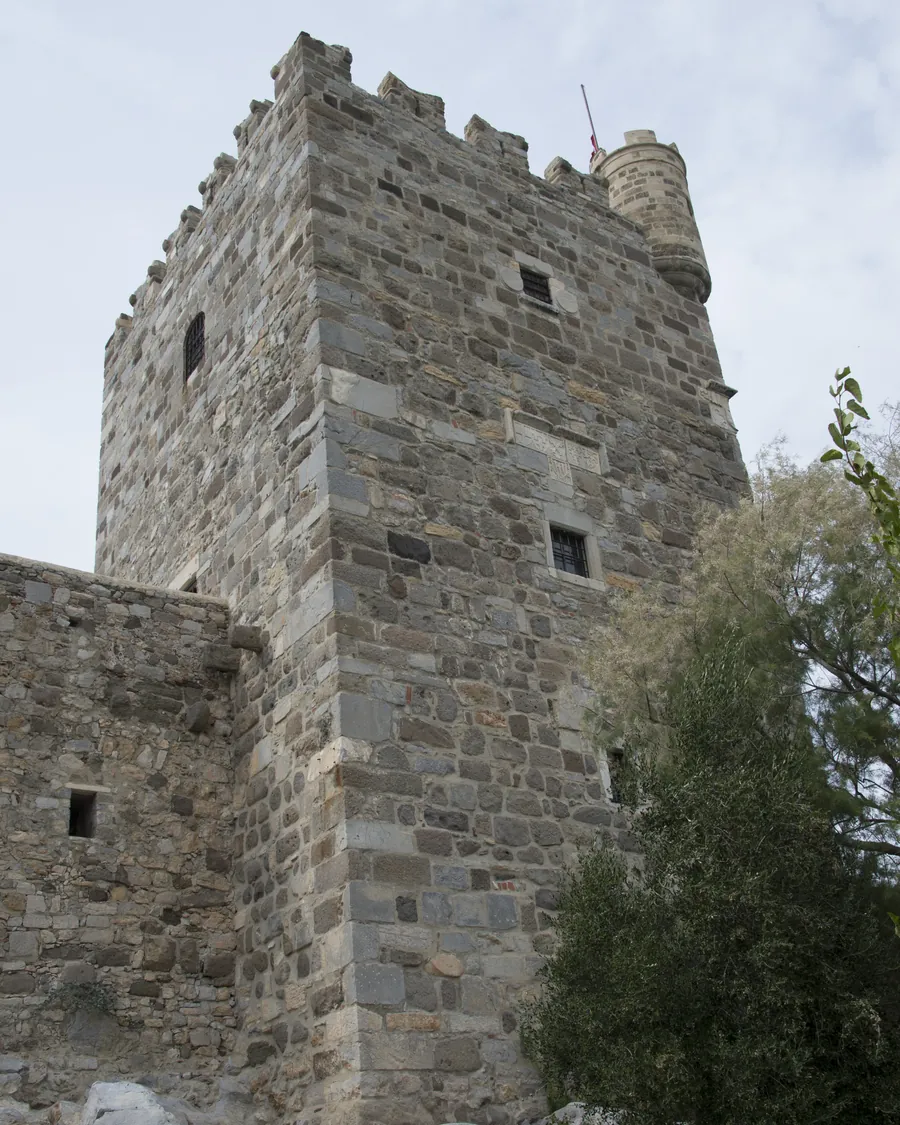 Ein quadratischer Steinturm mit Zinnen und einem kleinen Eckt&uuml;rmchen, von unten vor hellem Himmel gesehen, mit Felsen und einem Baum am Fu&szlig;