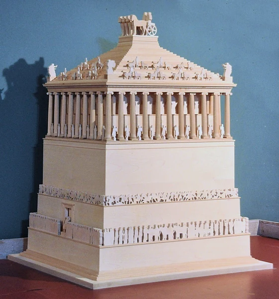Ein helles Holzmodell eines hohen Grabmals mit gestuftem Sockel, einer S&auml;ulenreihe, einem Pyramidendach und einem Wagen darauf, vor einer blauen Wand