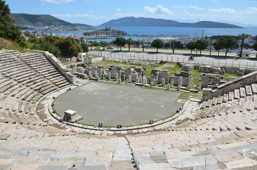 Ein steinernes Theater mit geschwungenen Sitzreihen um eine runde ebene Fl&auml;che, dahinter Ruinen und in der Ferne ein Hafen und H&uuml;gel
