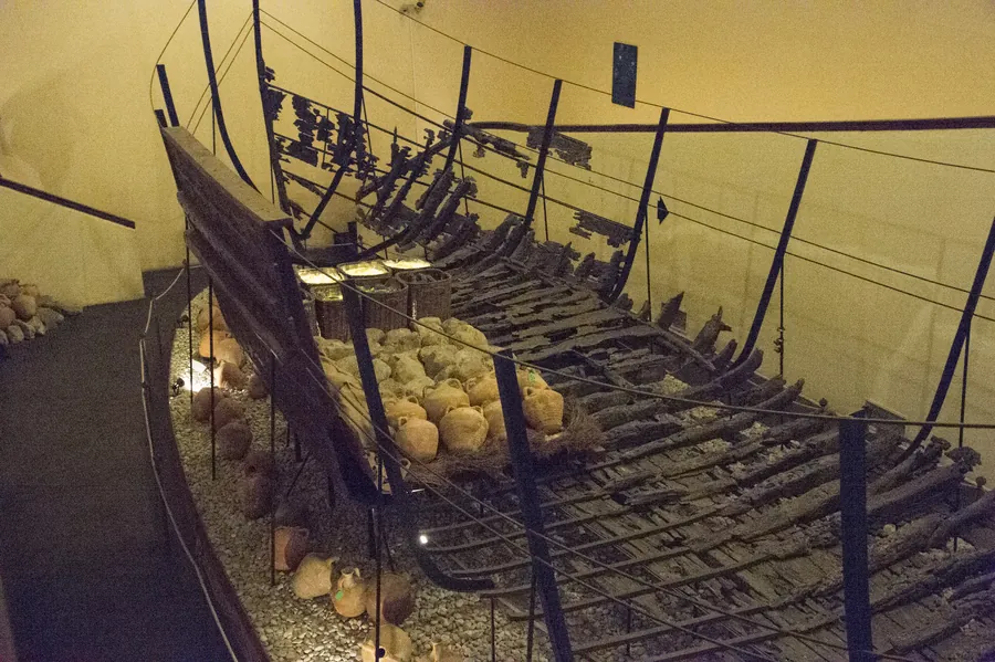 Die dunklen, gebogenen H&ouml;lzer eines Schiffswracks in einem Museumssaal, im Rumpf liegen Reihen heller Tongef&auml;&szlig;e
