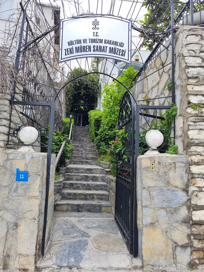 Eine Steintreppe f&uuml;hrt hinter einem Eisentor zwischen gr&uuml;nen Pflanzen hinauf, &uuml;ber dem Tor ein wei&szlig;es Schild mit der Aufschrift Zeki M&uuml;ren Sanat M&uuml;zesi