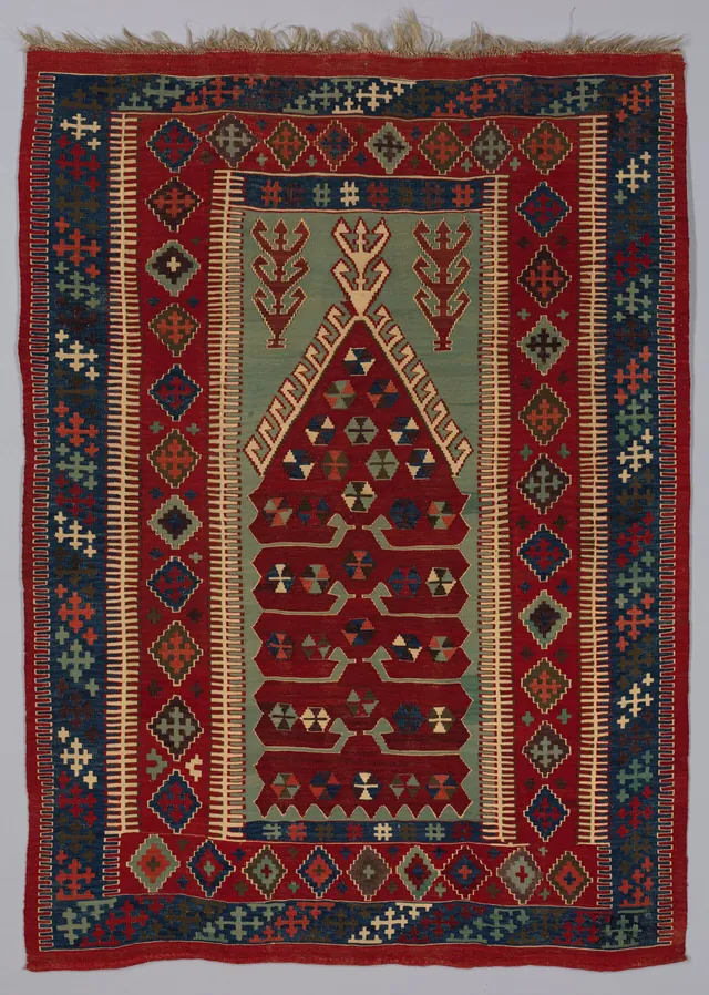 La parte superiore di un kilim a tessitura piana con campo rosso, ampie bordure blu a piccole croci e rombi, un riquadro centrale verde con una nicchia a punta e una frangia in alto