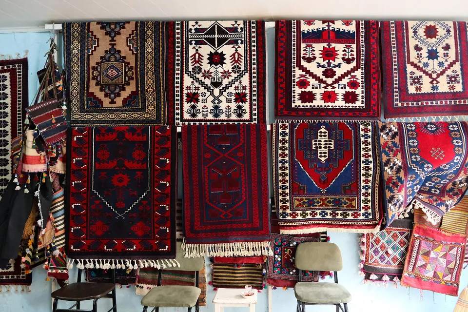 Una parete con due file di tappeti a pelo rosso scuro, blu marino e crema, decorati con medaglioni a gradini e nicchie di preghiera, e piccole borse tessute accanto