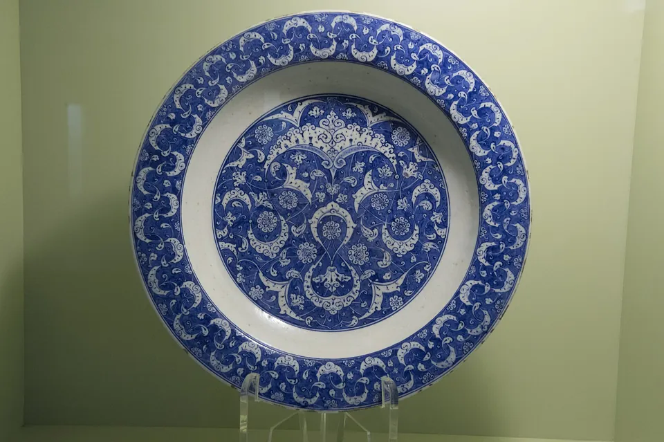Un grande piatto con un’ampia bordura blu a foglie ricurve e un medaglione blu di volute e rosette su fondo bianco, su un supporto trasparente in una vetrina illuminata