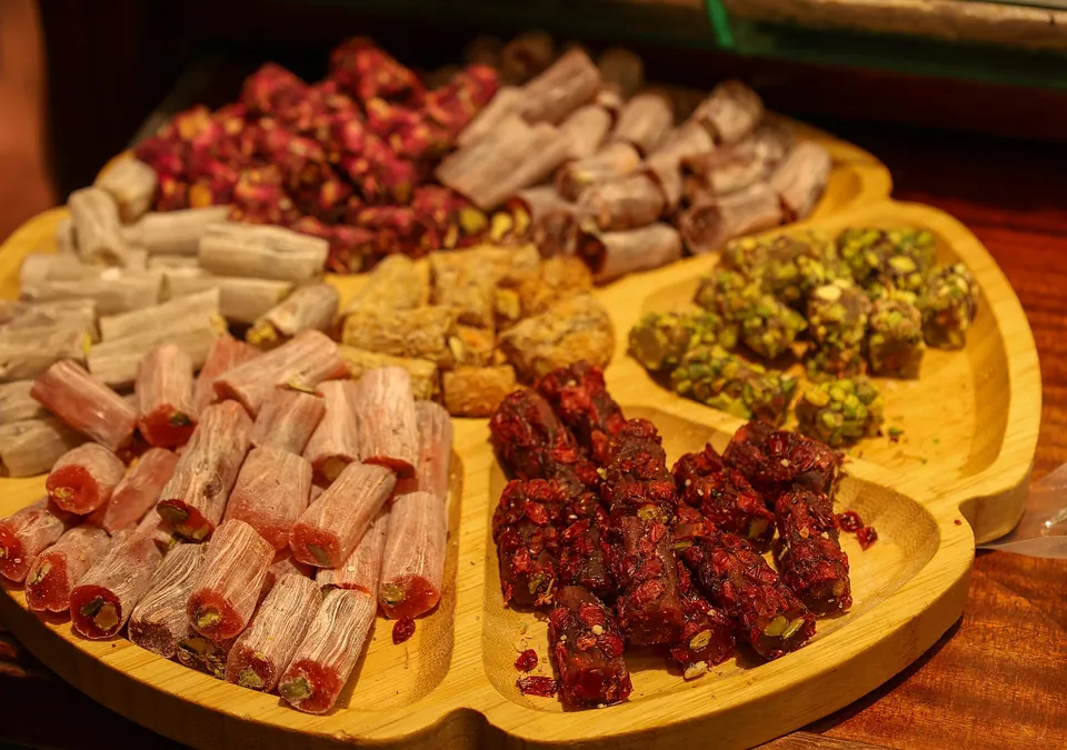 Un vassoio di legno dal bordo sagomato, diviso in scomparti con delizie turche: pezzi arrotolati ricoperti di petali di rosa, bastoncini chiari, rotoli al pistacchio e pezzi scuri con frutta