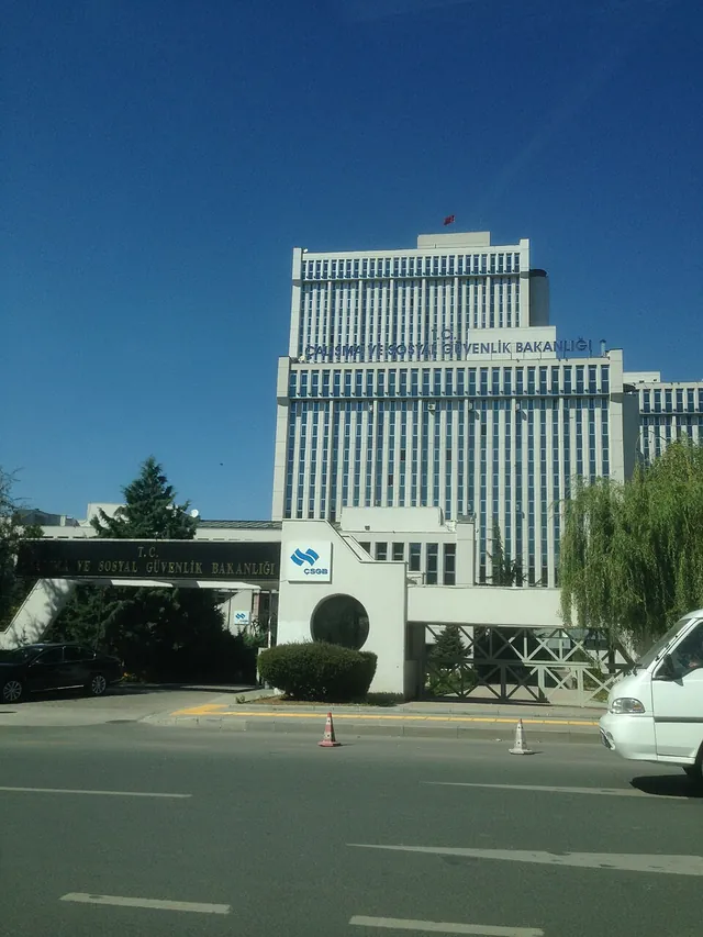 A tall white office tower of the labour ministry behind a gate and a low entrance building in Ankara, under a blue sky