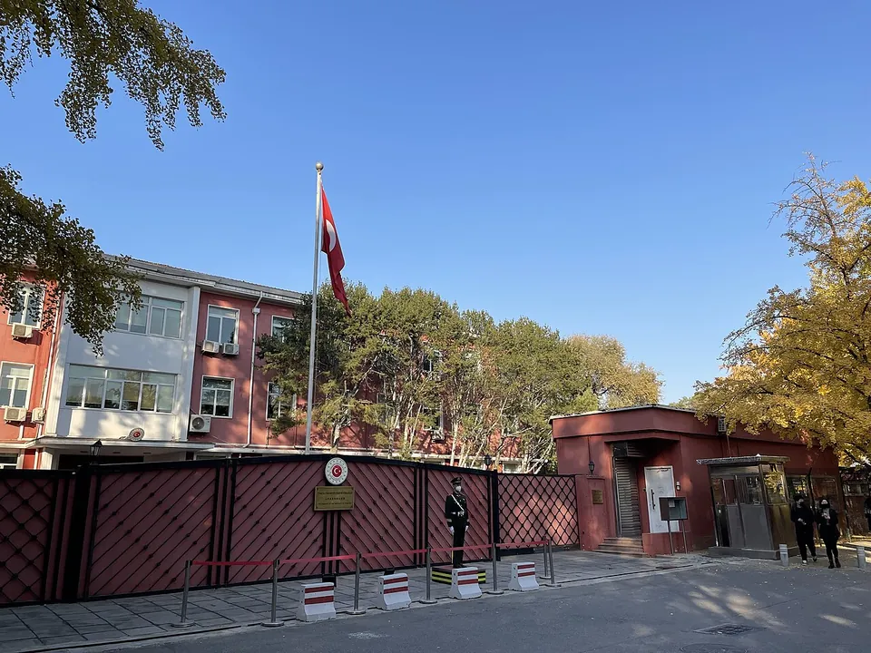 The red-brown wall and gate of the Turkish embassy in Beijing with the Turkish flag on a pole, a guard at the gate and autumn trees
