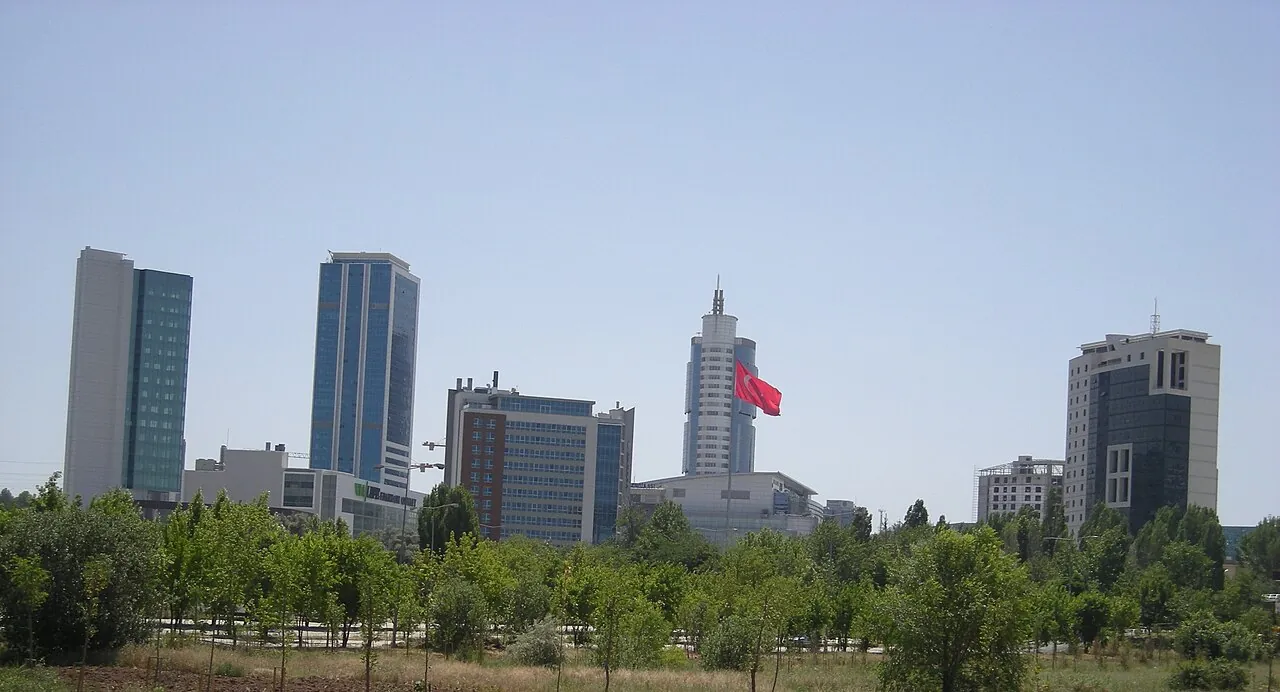 High-rise office towers of the Söğütözü district in Ankara over a park, one with a Turkish flag on its roof, under a hazy sky