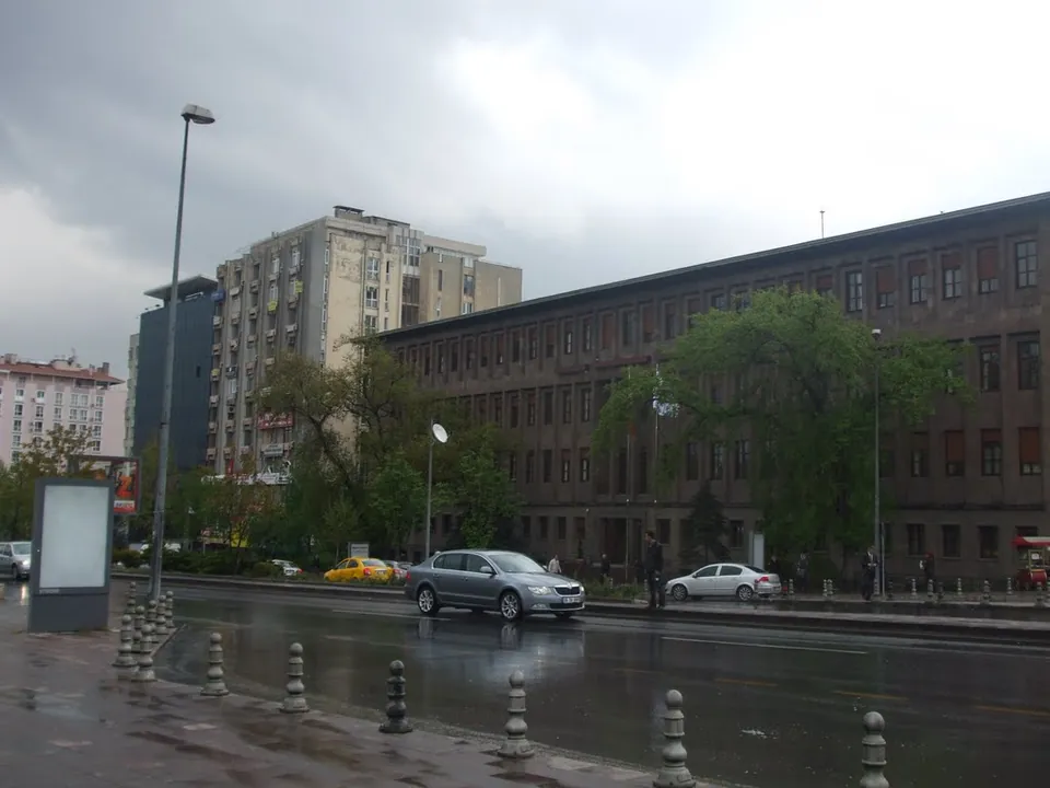 A long brick ministry building and an apartment block on a wide wet avenue in Kızılay, Ankara, with cars and street lamps