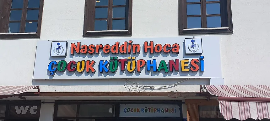 واجهة مبنى بيضاء، عليها لافتة طويلة زرقاء وبيضاء بأحرف ملوّنة تحمل اسم Nasreddin Hoca Çocuk Kütüphanesi، مع شعارين دائريين وثلاث نوافذ علوية