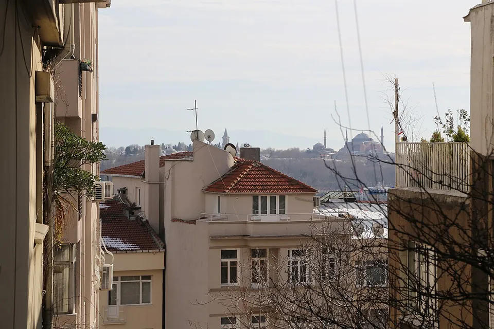 Daken van oude flatgebouwen in Istanbul met antennes en bovengrondse draden, met ver daarachter de koepel en minaretten van een moskee
