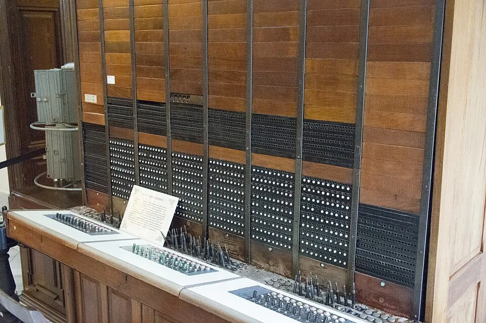 Een wandvullend houten telefoonschakelbord met rijen zwarte aansluitingen en een tafel met stekkers ervoor, in een museum