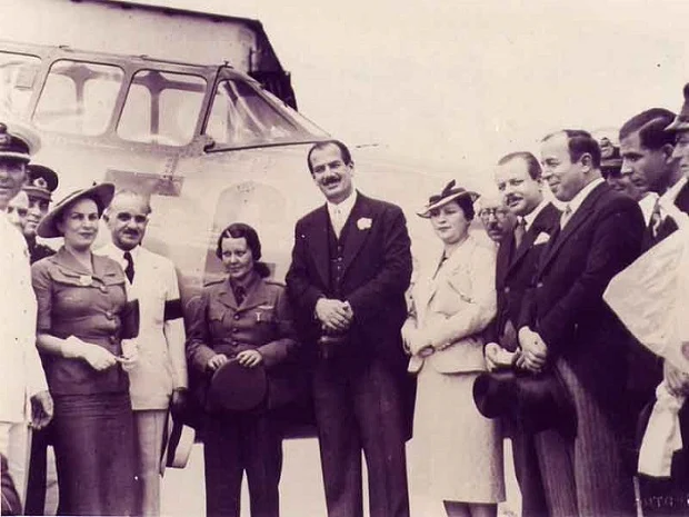 Fotografie: o femeie &icirc;n uniforma forțelor aeriene, cu chipiul &icirc;n m&acirc;nă, stă l&acirc;ngă un bărbat &icirc;n costum &icirc;nchis la culoare, &icirc;n fața cabinei vitrate a unui avion, &icirc;nconjurați de bărbați și femei cu pălării