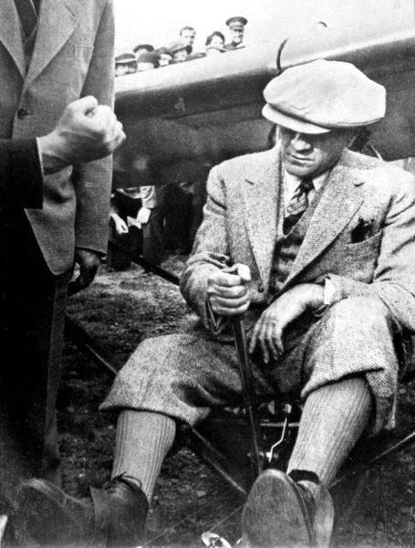 Fotografie: un bărbat cu șapcă și costum de tweed cu pantaloni p&acirc;nă la genunchi stă pe cadrul unui avion mic și ține manșa; &icirc;n spatele lui se văd o mulțime de oameni și o aripă