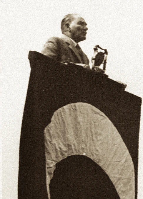 Fotografie: Atat&uuml;rk vorbește la un pupitru cu microfon; peste partea din față a pupitrului at&acirc;rnă o p&acirc;nză &icirc;nchisă la culoare, cu o semilună deschisă la culoare