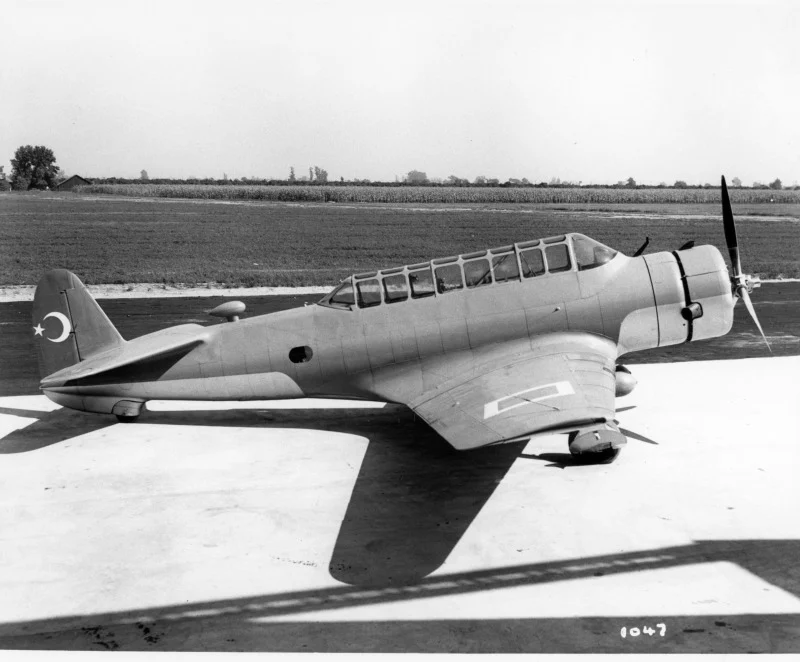 Fotografie: un avion monomotor cu aripă joasă, cabină lungă vitrată și carenaje fixe ale roților, cu o semilună și o stea pe coadă, pe o platformă de beton, cu un c&acirc;mp &icirc;n spate