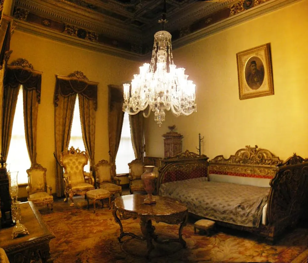A room in Dolmabahçe Palace with a gilded ceiling, a crystal chandelier, tall curtained windows and a daybed