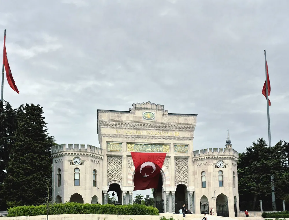 The gate of Istanbul University: a pale stone gateway with towers and clocks and a large Turkish flag hanging in the arch