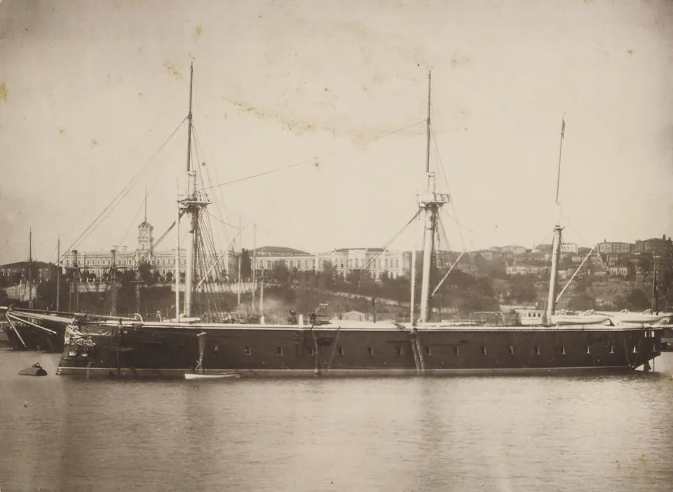 Sepia photograph of a dark ironclad warship with three masts at anchor, with buildings on a hill behind it