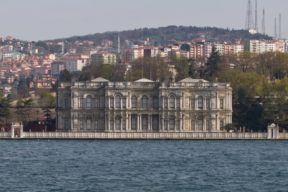 The two-storey stone façade of Beylerbeyi Palace on the Asian shore behind a low sea wall, with houses climbing the hill behind it