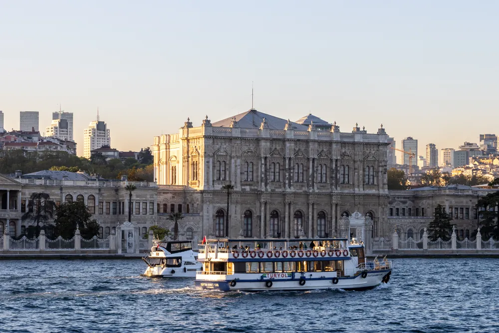 The sea façade of Dolmabahçe Palace seen from a boat on the Bosphorus, with a ferry in front and modern towers in the distance