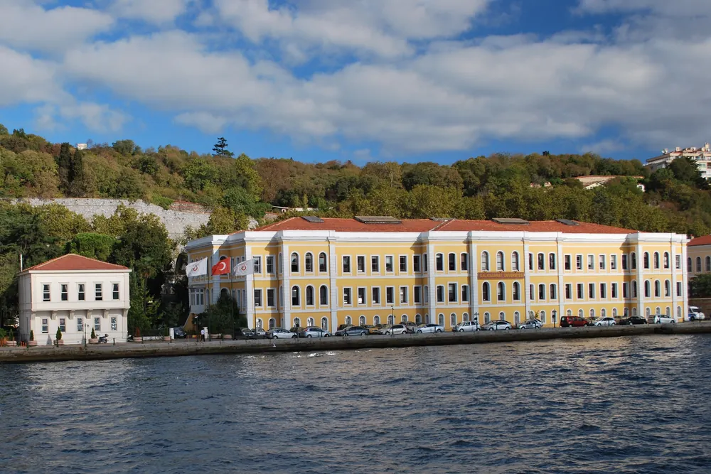 A long yellow two-storey building with a red-tiled roof on the Ortaköy shore, seen from the water, with a Turkish flag on a tall pole at the left
