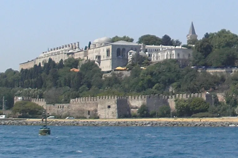 Topkapı Palace on its headland, seen from the water: long pale buildings with chimneys and a pointed tower above a battlemented sea wall and a green slope