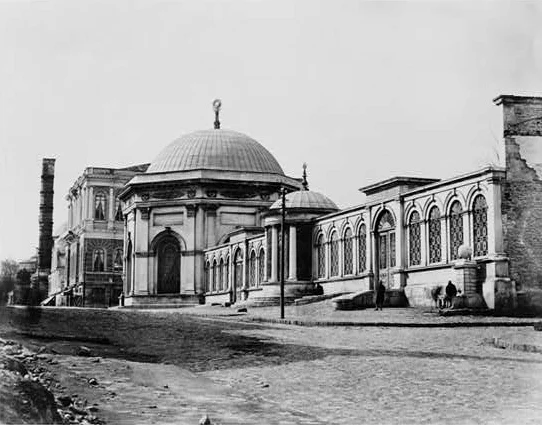 Fotograf&iacute;a antigua de un edificio funerario con c&uacute;pula y ventanas de arco junto a una calle, con una farola delante y una segunda c&uacute;pula peque&ntilde;a a un lado