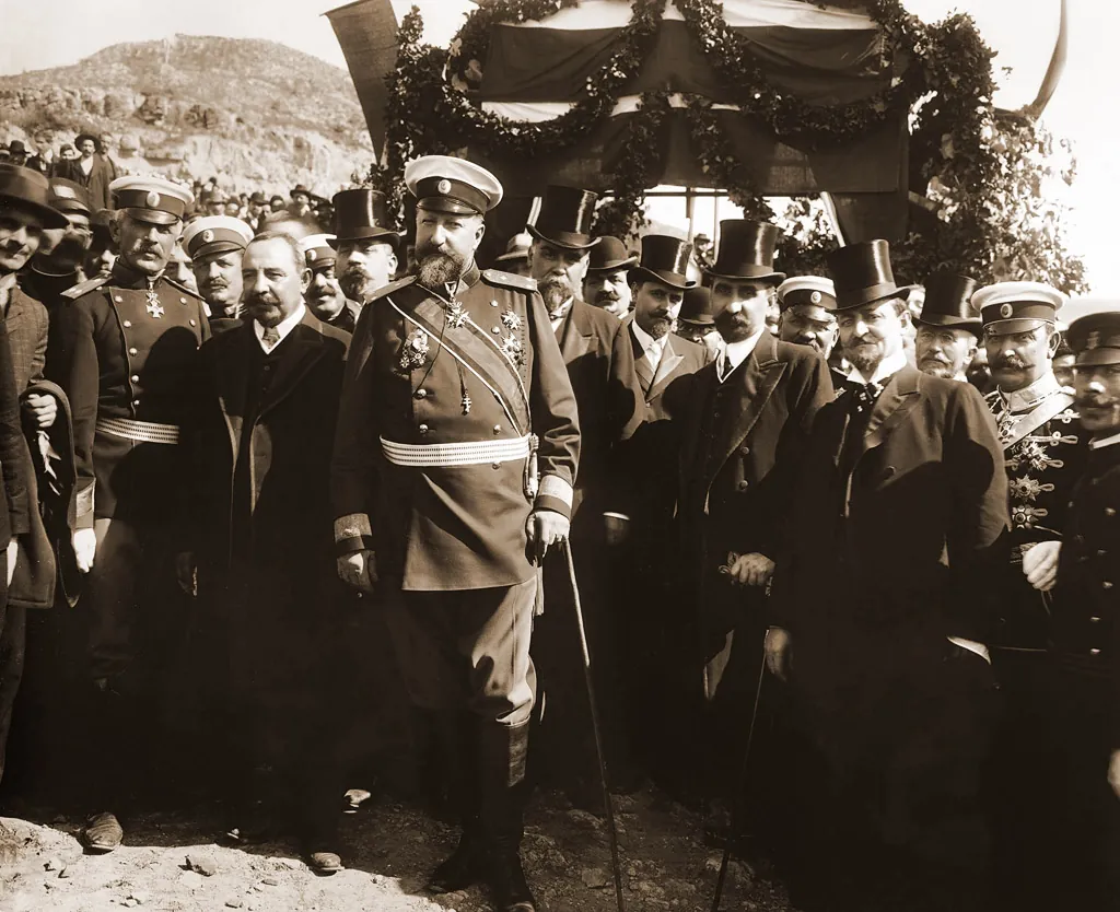 Sepiafoto: F&uuml;rst Ferdinand mit Bart, Milit&auml;rmantel, M&uuml;tze und wei&szlig;em Koppel, vor einer Menge von M&auml;nnern mit Zylindern und in Uniformen unter einem mit Kr&auml;nzen und Flaggen geschm&uuml;ckten Bogen
