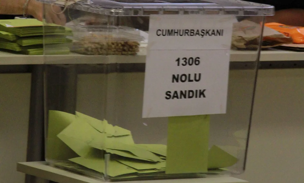 Eine durchsichtige Wahlurne mit einem weißen Papierschild mit der Aufschrift „Cumhurbaşkanı“ und „1306 nolu sandık“ und vielen olivgrünen Wahlumschlägen darin