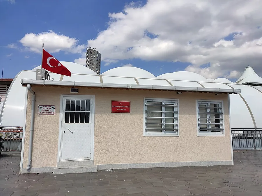 Ein kleines beiges eingeschossiges Gebäude mit weißer Tür, vergitterten Fenstern, einem roten Schild mit dem Namen des Büros eines Stadtviertelvorstehers und einer türkischen Fahne auf dem Dach; dahinter weiße Kuppeldächer unter blauem Himmel