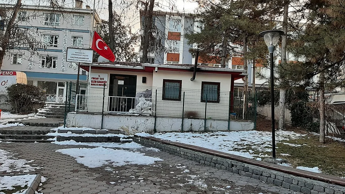Ein kleines weißes eingeschossiges Gebäude mit rotem Dachrand, einer türkischen Fahne an einem Mast und dem Schild des Büros eines Stadtviertelvorstehers; auf dem gepflasterten Weg davor liegt Schnee