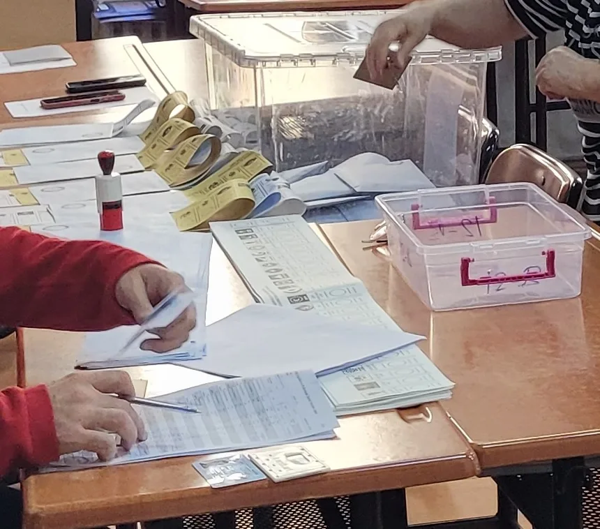 Zwei Tische mit einer durchsichtigen Wahlurne, Rollen gelber Aufkleber, einem Stempel mit rotem Griff, Stimmzetteln, einer Wählerliste und einer durchsichtigen Kunststoffablage mit rosa Griffen; dazu die Hände von Mitgliedern des Wahlvorstands und eines Wählers, der einen Wahlumschlag über die Urne hält