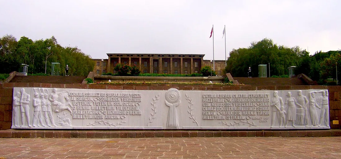 Eine lange weiße Marmorreliefwand mit gemeißelten Figuren und Textzeilen vor terrassenförmigen Stufen; dahinter auf dem Hügel das lange Gebäude der Nationalversammlung mit zwei Fahnenmasten unter bewölktem Himmel