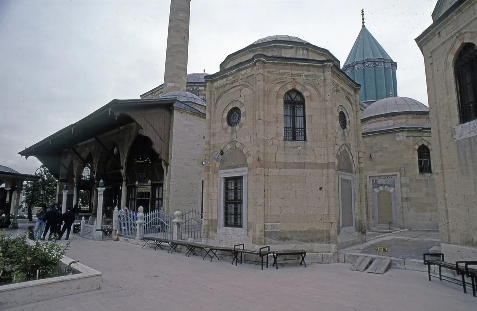 O p&aacute;tio do Museu Mevlana: um pr&eacute;dio de pedra com janelas em arco e uma c&uacute;pula baixa, um alpendre de madeira &agrave; esquerda e a c&uacute;pula c&ocirc;nica turquesa atr&aacute;s
