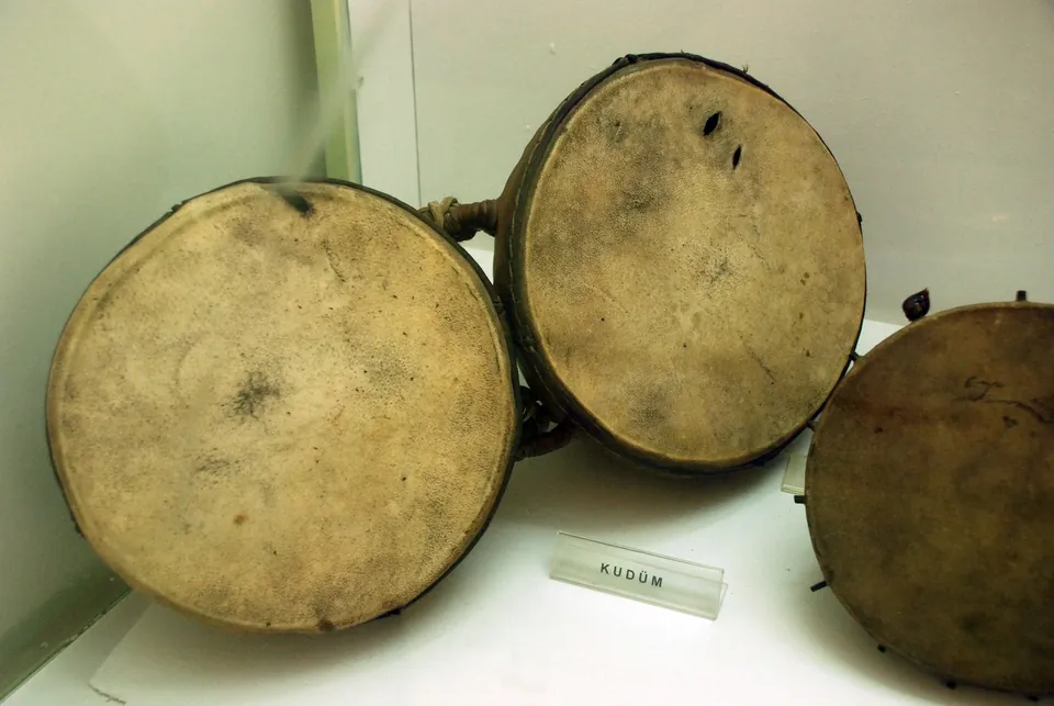Tr&ecirc;s tambores redondos com membranas de pele numa prateleira de museu, com uma pequena etiqueta com a inscri&ccedil;&atilde;o KUD&Uuml;M